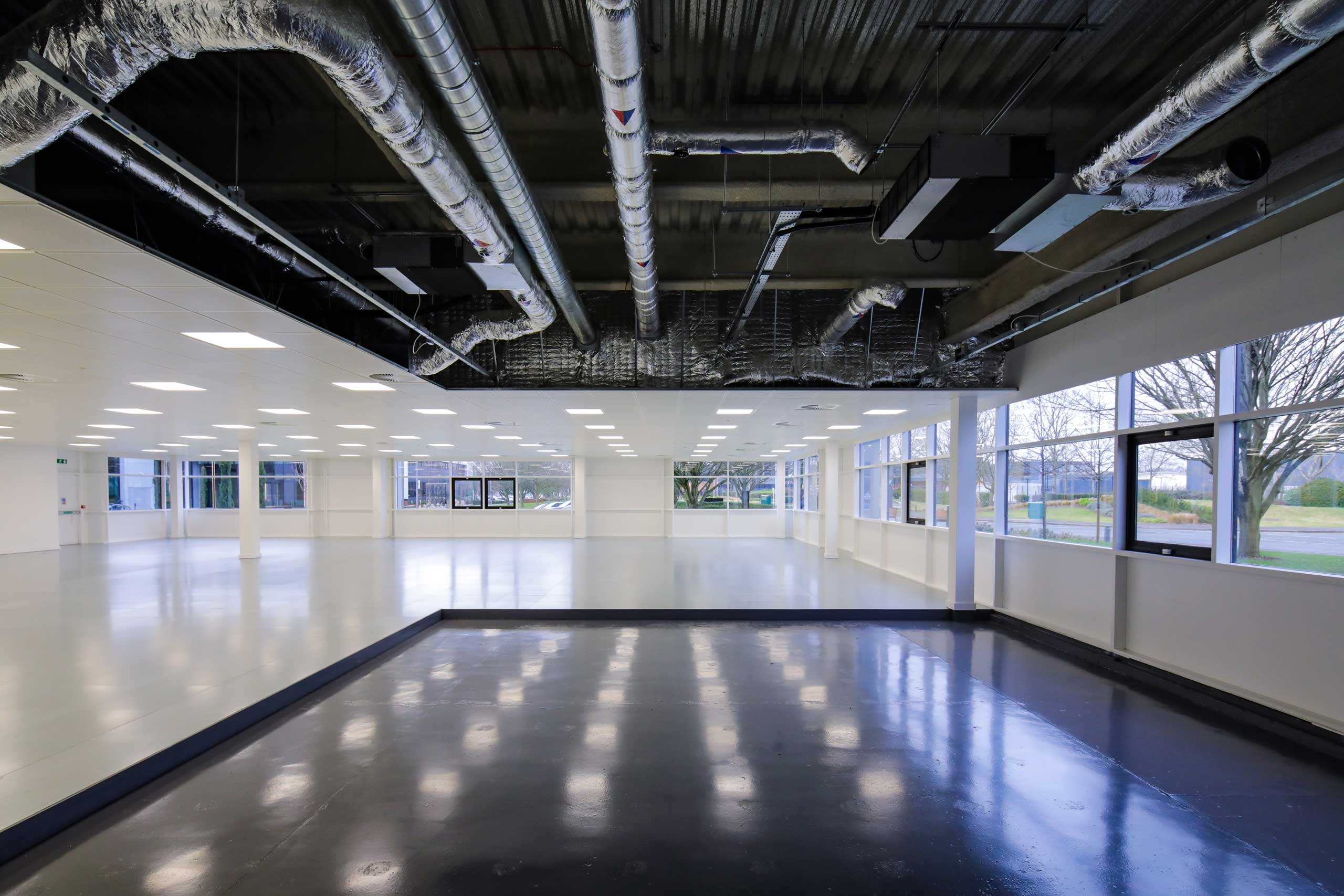 Empty industrial-style office space with exposed ceiling ducts and large windows on two sides.