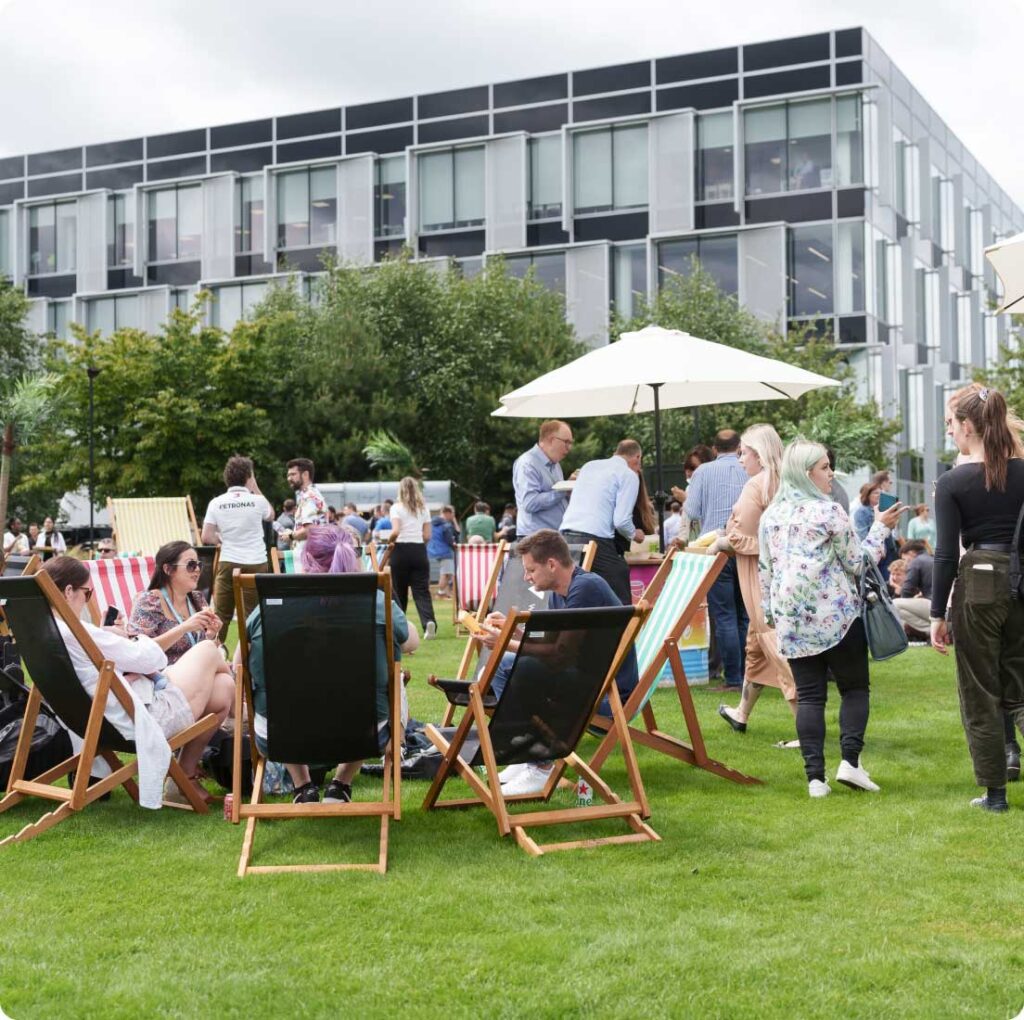 A group of people are sitting on deck chairs and standing on a grassy lawn in front of a modern building, socializing and interacting under an overcast sky.