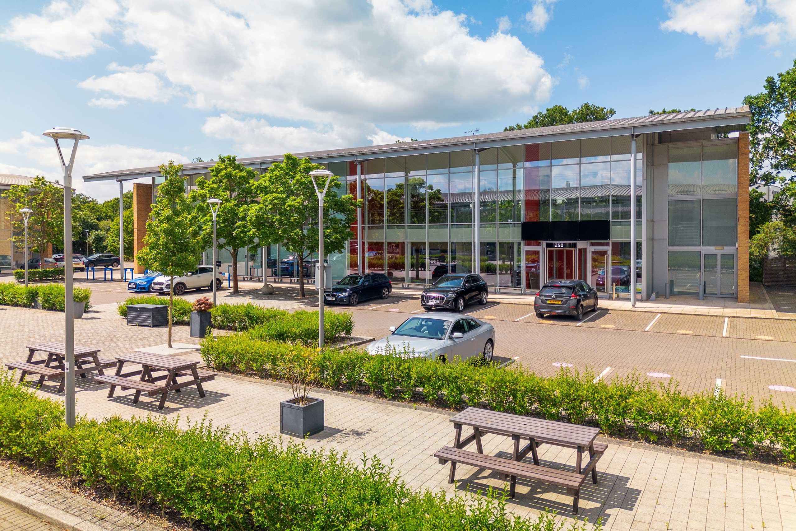 Modern glass-fronted building with parked cars and trees in the foreground. Three picnic tables are on a paved area with some plants and light poles under a partly cloudy sky.